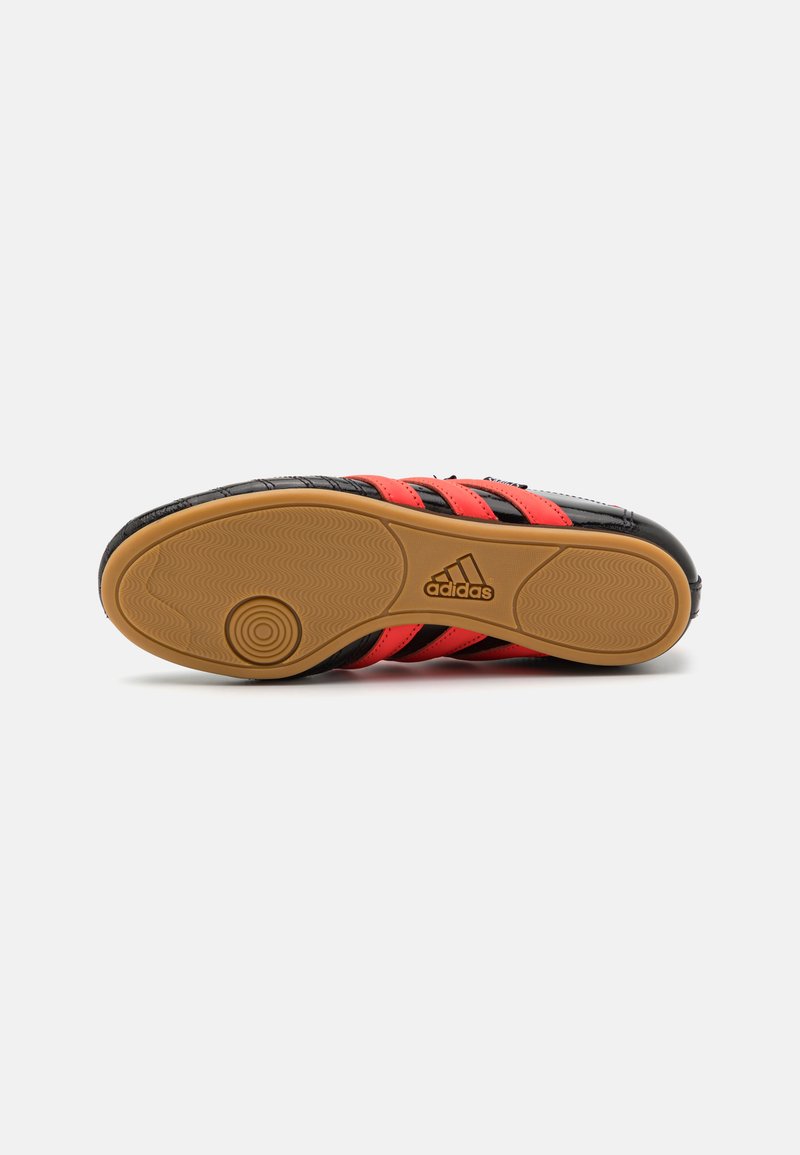 Zapatilla deportiva negra con detalles a rayas rojas y negras, con una suela de goma texturizada y un logo distintivo. Diseñada para el rendimiento y la tracción.