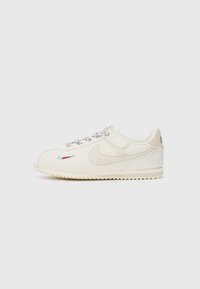 Off-white lav topp sneaker med beige Nike swoosh, flerfargede snører og brodert mini swoosh nær tåen på hvit bakgrunn.