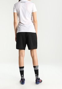 Bílá krátká košile s krátkým rukávem, černé šortky, vzorované šedé a černé ponožky, modré kopačky, pohled zezadu. Jednoduchý, atletický design.