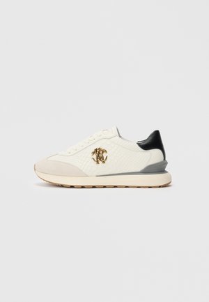 Vit lågtopp sneakers med texturerad sida, beige tåkappa, svart häl, grå mellansoffaccent och guld logotyp med sammanlänkande design på sidan.