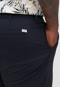 Los pantalones azul marino presentan un bolsillo trasero con una etiqueta blanca que dice "JACK & JONES". La tela es suave, con un brillo sutil y un corte estructurado.