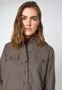 Camicia marrone a quadri con due tasche sul petto, chiusura con bottoni e texture morbida. Indossata sopra un dolcevita più scuro.