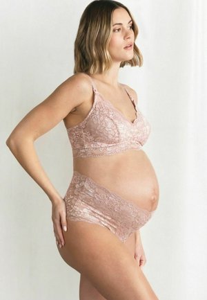 Pregnant woman wearing matching pink lace maternity bra and high-waisted underwear, standing in profile against a light background.