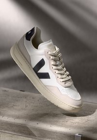 White leather sneaker with dark blue accents, beige suede details, perforated toe, flat laces, and a textured cream rubber sole.