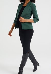 Grön åtsittande kavaj med en strukturerad design, med trekvartsärmar. Kombinerad med en svart ribbad klänning och knähöga svarta stövlar.