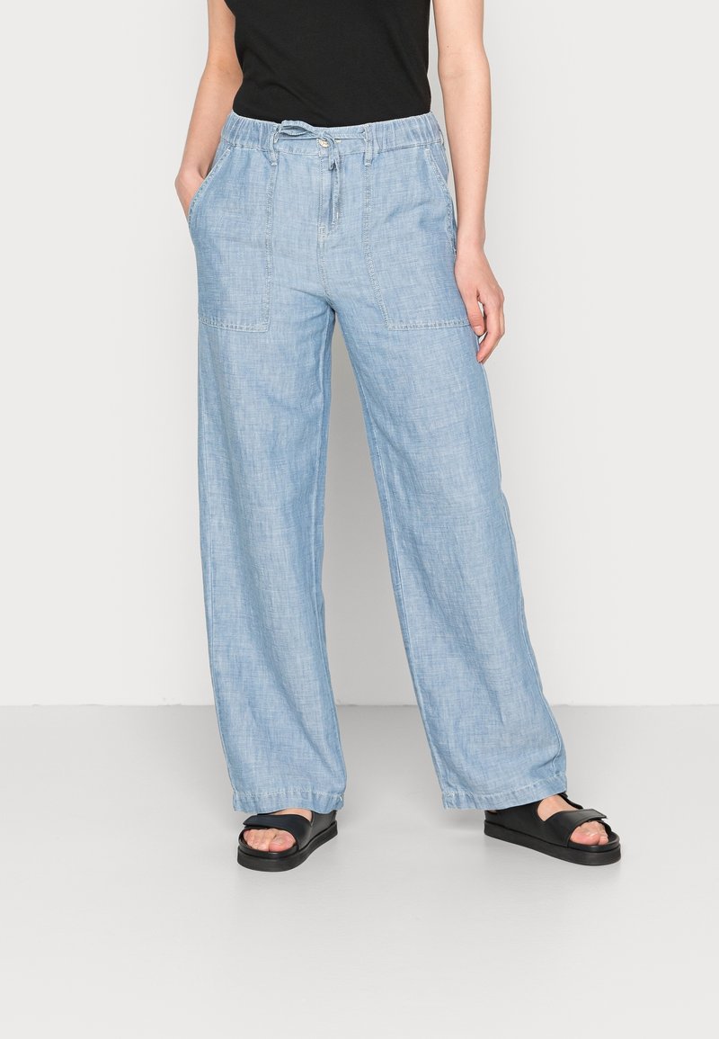 Persoon in lichtblauwe wijde linnen broek met elastische tailleband en zwarte slip-ons, staand tegen een effen witte achtergrond.