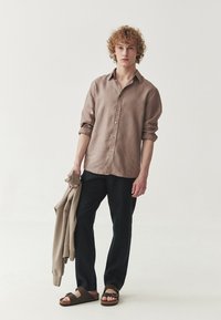 Camisa de lino marrón claro con botones, pantalones oscuros azul marino, sudadera beige caida sobre el brazo y sandalias marrones con tiras.