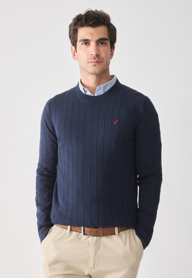 Pull en tricot côtelé marine avec un col rond et des manches longues, arborant un petit logo rouge sur la poitrine, associé à une chemise à col bleu clair.