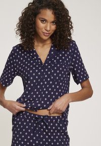 Laivastonsininen pyjama-setti, jossa on lyhyet hihat, napillinen paita ja naruhelmaiset housut, joissa on valkoisia timanttikuosisia kuvioita. Pehmeä kangastuntuma.
