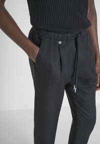 Black linen trousers with a tapered design, elastic waistband, and drawstring. Features side pockets and subtle metallic hardware accents.