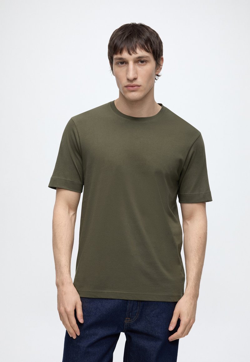 Jeune homme aux cheveux courts et foncés, portant un t-shirt vert olive à manches courtes et un jean bleu foncé, debout devant un fond clair uni.
