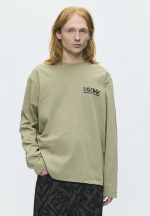 Jeune personne aux longs cheveux roux portant une chemise oversize à manches longues vert olive clair avec un texte noir et un pantalon foncé à motifs, debout devant un fond blanc.