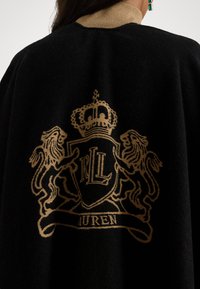 Melnā apmetņa ar bežīgu apkakli, kuru rotā zelta izšuvums ar ģerboni, uz kura attēloti lauvas un kronis, apakšā ir redzams uzraksts "RLL" un "LAUREN".