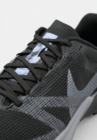 Baskets en maille noire avec une surface texturée, présentant des accents gris et un logo réfléchissant. Les lacets noirs assortis complètent le design.