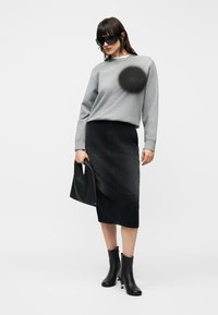 Γυναίκα που φοράει υπερμεγέθη μαύρα γυαλιά ηλίου, γκρι φούτερ, μαύρη μίντι φούστα, μαύρες μποτάκια μέχρι τον αστράγαλο, κρατώντας μικρή μαύρη τσάντα, στέκεται μπροστά σε λευκό φόντο.