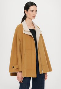 Cappotto a mantella in lana marrone con colletto in ecopelliccia crema, tasche laterali, chiusura con zip e dettagli in bottone sulle maniche. Texture liscia.