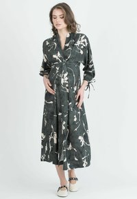 Langes, dunkelgrünes Kleid mit einem floralen Muster in Cremetönen. Weicher Stoff, Wickeldesign, gebundene Ärmel und ein fließender Rock. Eignet sich gut für Umstandsmode.