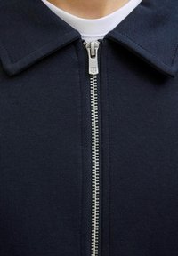 Jūrinės mėlynos zip-up striukės su smailiu apykakle ir struktūrizuota medžiaga. Savybės: sidabrinis užtrauktukas ir subtilus siuviniai palei apykaklę.