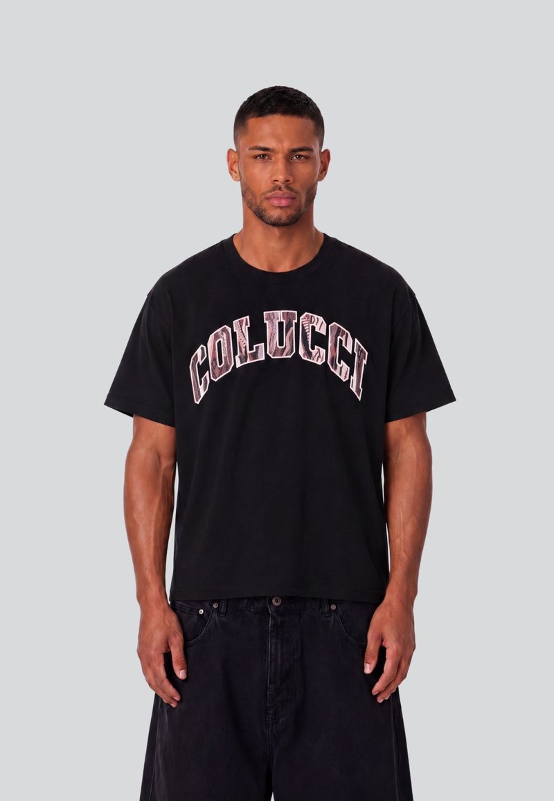 Άνδρας που φοράει μαύρο t-shirt Colucci και μαύρο παντελόνι, στέκεται μπροστά σε απλό γκρι φόντο με τα χέρια χαλαρά δίπλα στο σώμα.