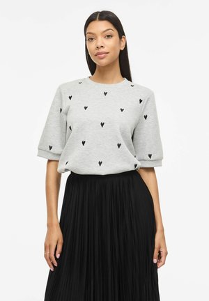Kvinde med langt mørkt hår iført lysegrå kortærmet top med sorte hjerteprint og sort højplisseret nederdel.