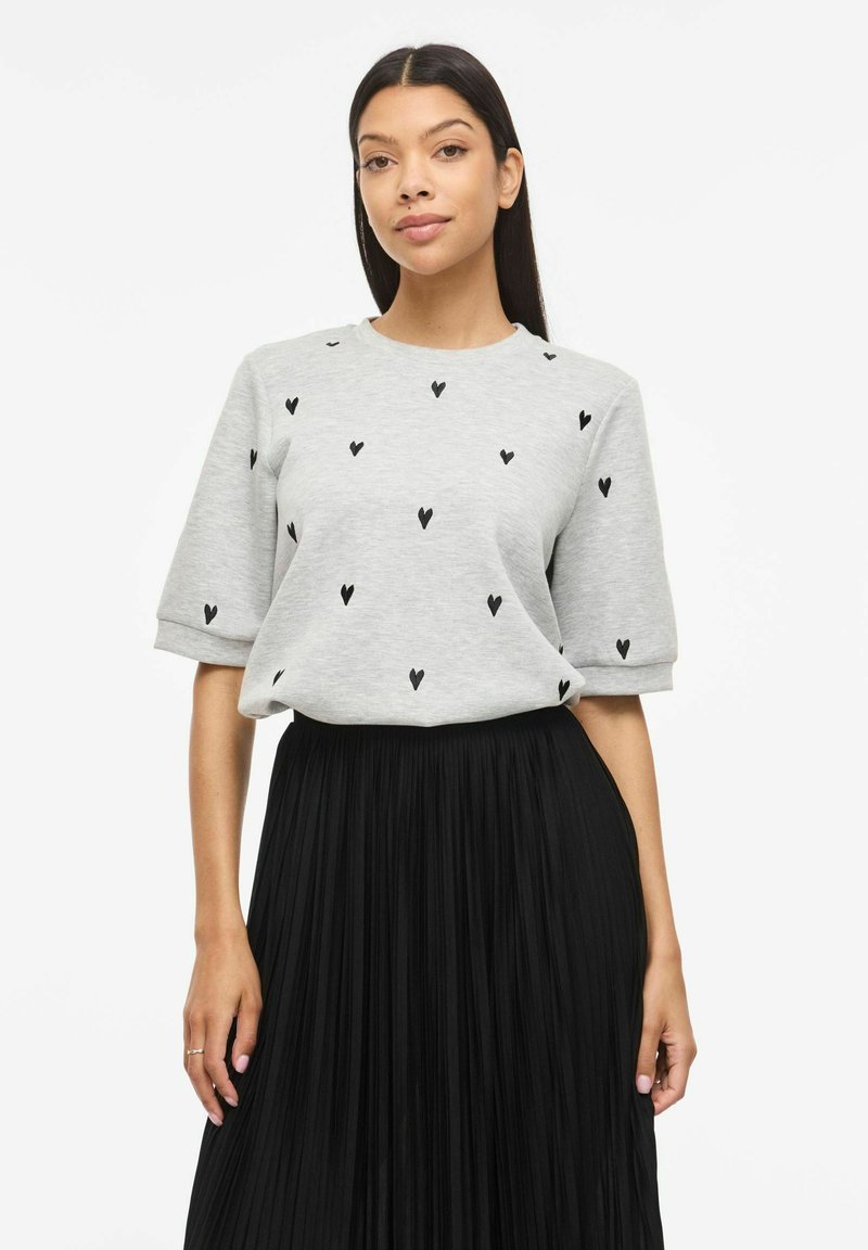 Kvinde med langt mørkt hår iført lysegrå kortærmet top med sorte hjerteprint og sort højplisseret nederdel.