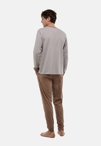 Chemise gris clair à manches longues avec des rayures texturées, associée à un pantalon de jogging beige avec des poignets élastiques. Le mannequin se tient pieds nus, les mains dans les poches.