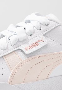 Vit lädersko med texturerad yta, mjuka rosa mockadetaljer, plana vita snören och en liten röd logotypetikett på sidan.