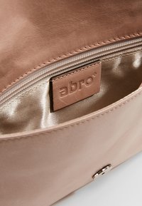 Intérieur d'un sac en cuir rose clair avec une doublure satinée, comportant une étiquette marron gaufrée avec "abro" et un détail en métal argenté de forme ronde.