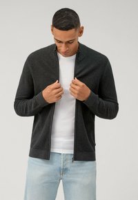 Grijze geribbelde rits cardigan met hoge kraag, gedragen over een wit shirt. Model houdt het kledingstuk vast, gecombineerd met lichtblauwe jeans.