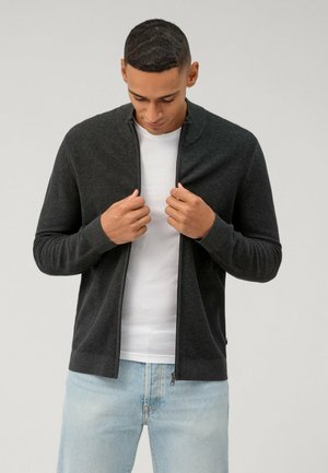 Grijze geribbelde rits cardigan met hoge kraag, gedragen over een wit shirt. Model houdt het kledingstuk vast, gecombineerd met lichtblauwe jeans.