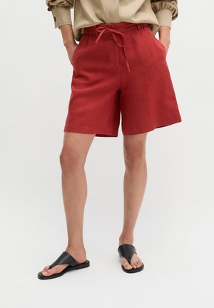 Osoba w czerwonych szortach do kolan ze sznurkiem w pasie i beżowej bluzce z długim rękawem, stoi z rękami w kieszeniach i czarnych sandałach.