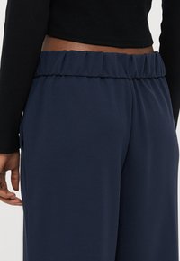 Person som har på seg mørkeblå elastiske bukser med strikk i livet og en svart, langermet crop top, med hånden avslappet ned langs siden.