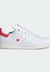 Sneaker in pelle bianca con linguetta rossa sul tallone, logo Adidas rosso e lacci bianchi su suola bianca testurizzata, su sfondo chiaro semplice.