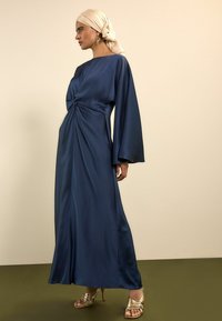 Tumesinist satiinist kleit, millel on vöökohal keeratud sõlme detail, pikad lehvikukujulised varrukad ja põrandani ulatuv siluett. Kullakarva rihmikandaga kontsakingad tekitavad kontrasti.