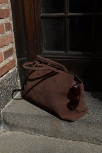 Brun mockaläder tote väska med två handtag, vilande på trappor, med akrylsolglasögon som ligger på sidan. Enkelt design, inga synliga logotyper.
