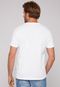 Camiseta de algodón blanca de mangas cortas, cuello redondo y un pequeño logo impreso en la parte trasera. Combinada con pantalones vaqueros de mezclilla azul.