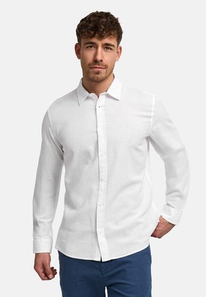 Mand med kort krøllet hår iført en langærmet hvid skjorte med knapper og blå bukser, der står mod en ensfarvet baggrund.