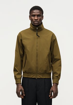 Férfi olívazöld cipzáras dzsekiben, magas gallérral és elasztikus mandzsettákkal, fekete nadrággal párosítva, egy egyszínű háttér előtt áll.