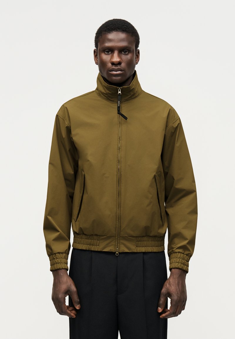 Férfi olívazöld cipzáras dzsekiben, magas gallérral és elasztikus mandzsettákkal, fekete nadrággal párosítva, egy egyszínű háttér előtt áll.