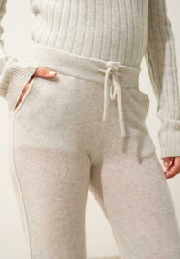 Světlé šedé kašmírové tepláky s pružným pasem, žebrovanými manžetami a jemnou texturou. Viditelné boční kapsy a jemné barevné variace.