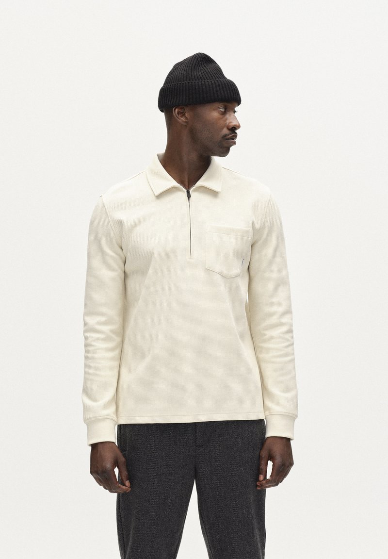 Világos krém színű, hosszú ujjú póló, texturált anyagból, cipzáras nyakkal és mellzsebből. Sötétszürke texturált nadrággal és fekete sapkával párosítva.