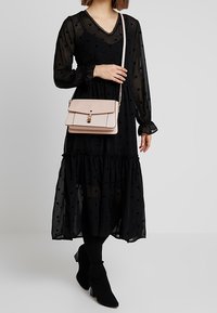 Abito maxi nero a pois con maniche lunghe e orlo arricciato, abbinato a una borsa a tracolla in pelle rosa chiaro con chiusura dorata e tracolla a catena.