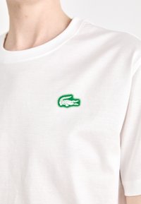 Biela bavlnená tričko s okrúhlym výstrihom, s malým zeleným vyšitým logom krokodíla na ľavej strane hrude. Krátke rukávy.