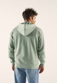 Felpa verde menta con cappuccio, dal taglio morbido, maniche lunghe e tasca a marsupio, abbinata a jeans blu in denim.