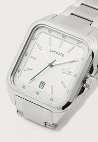 Kvadrātveida sudraba rokas pulkstenis ar baltu ciparnīcu, zaļiem akcentiem, datuma rādītāju 6 stundās un Lacoste logotipu ciparnīcas labajā pusē.