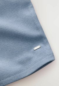 Sininen tekstiilipinta, jossa on pieni hopeinen tarra, jossa lukee "DOWNTIME", kiinnitetty helmaan. Reunat viimeistelty yksinkertaisella tikillä.
