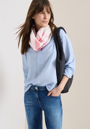 Vrouw met een lichtblauw bloesje, een roze-witte sjaal, een blauwe spijkerbroek en een zwarte gewatteerde tas over één schouder.