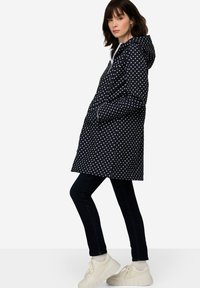 Vesimäräntä tummansininen pilkkukuosinen takki hupun kanssa, valkoiset yksityiskohdat, sivutaskut ja rento leikkaus, yhdistettynä tummiin farkkuihin ja näyttäviin valkoisiin kenkiin.