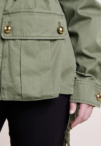 Veste verte avec poche à rabat boutonnée et boutons dorés, portée sur un pantalon noir, main détendue sur le côté.