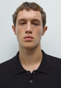 Jonge man met kort bruin haar in een zwart geribd poloshirt tegen een effen lichte achtergrond, kijkend recht vooruit.